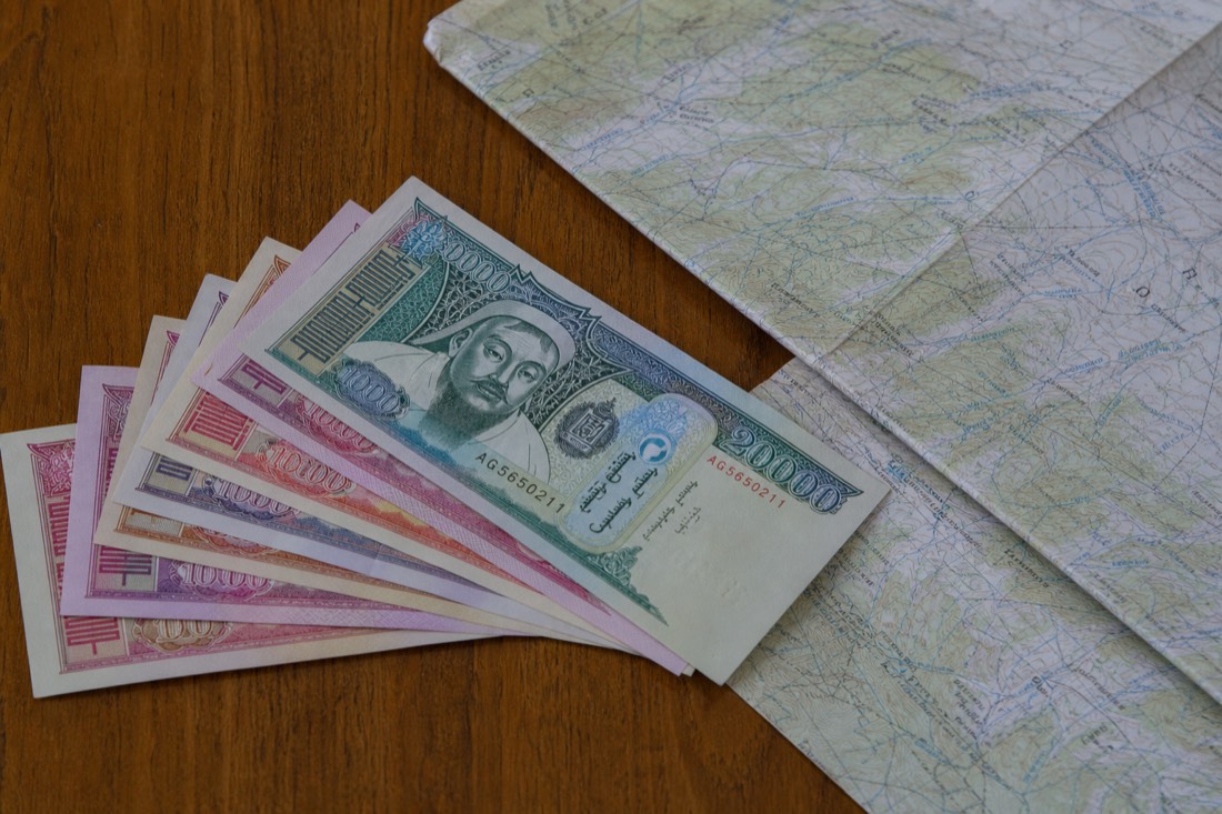 Mongolian tögrög banknotes fanned out on a wooden table beside a folded map of the Mongolian steppe, the cash visitors use to pay entrance and vehicle fees at the Hustai park gate